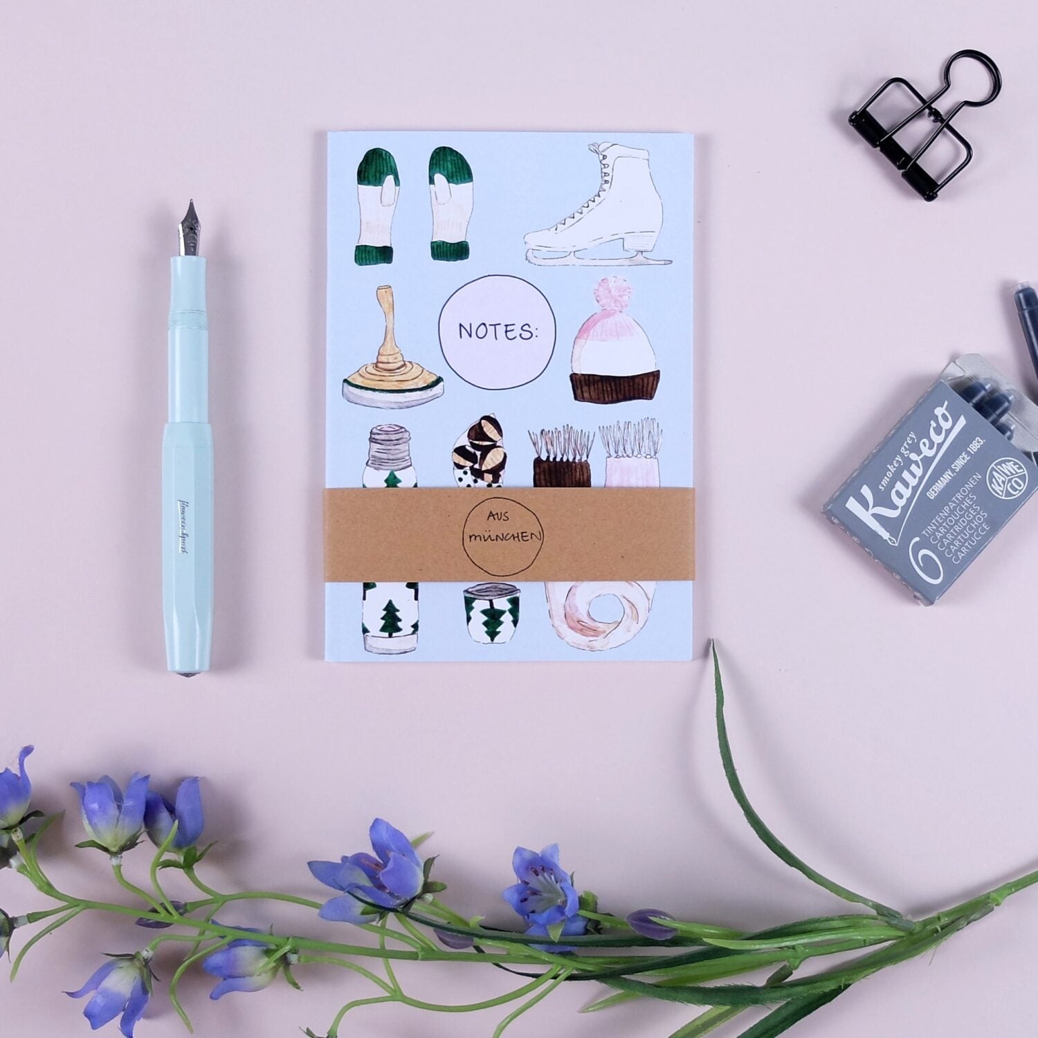 Notizbuch mit Wintermotiven, blauer Füllfederhalter links, Tintenpatronen rechts, blaue Blumen unten auf rosa Hintergrund.