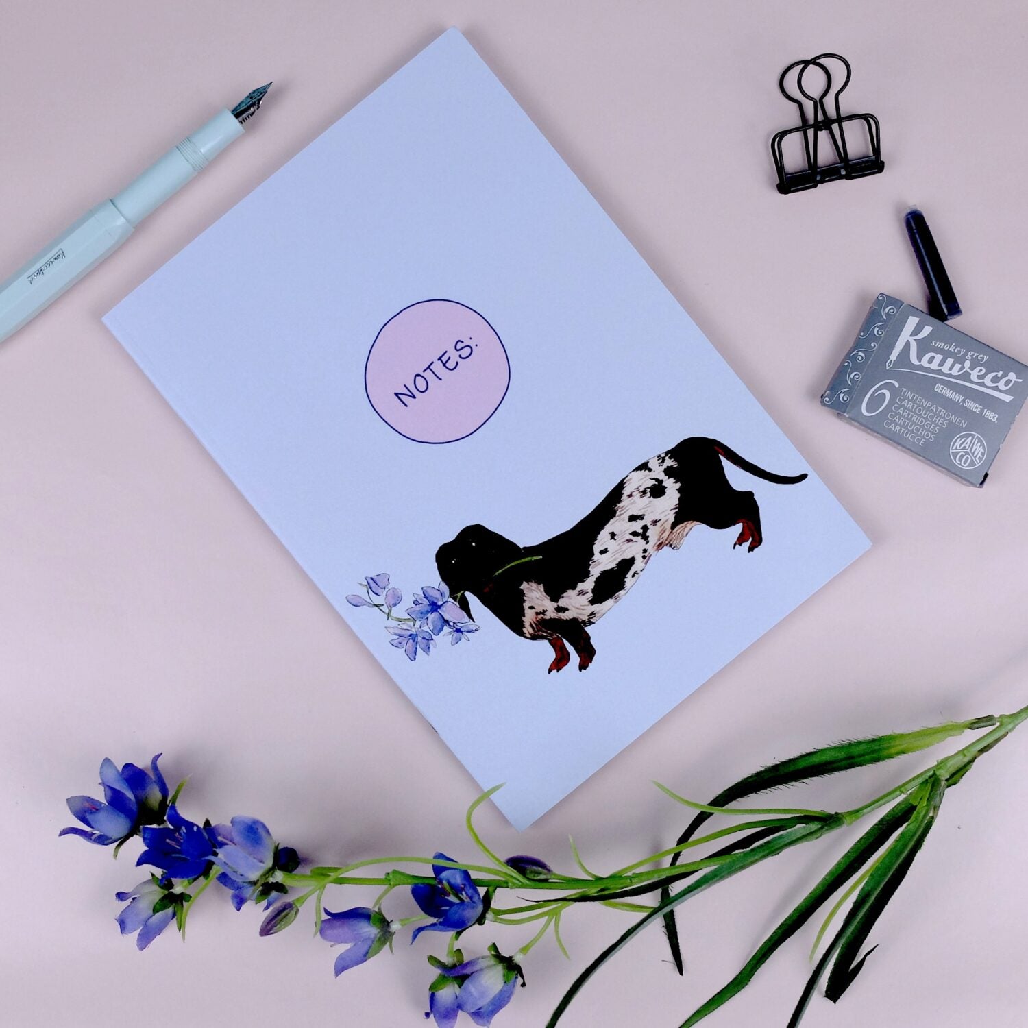 Notizbuch mit Dackelmotiv, Füllfederhalter, Tintenpatronenbox, Klammer und blauen Blumen auf rosa Hintergrund.