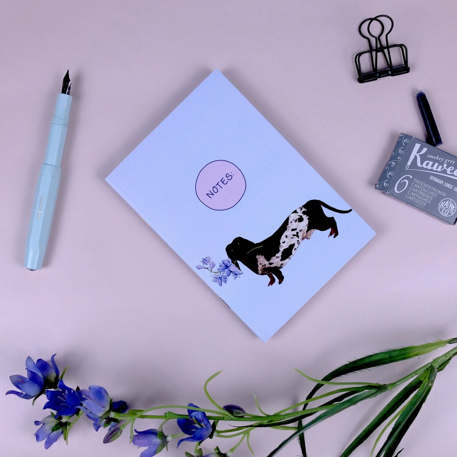 Notizbuch mit Hundemotiv, Füllfederhalter, Blumen und Büroklammern auf rosa Hintergrund.