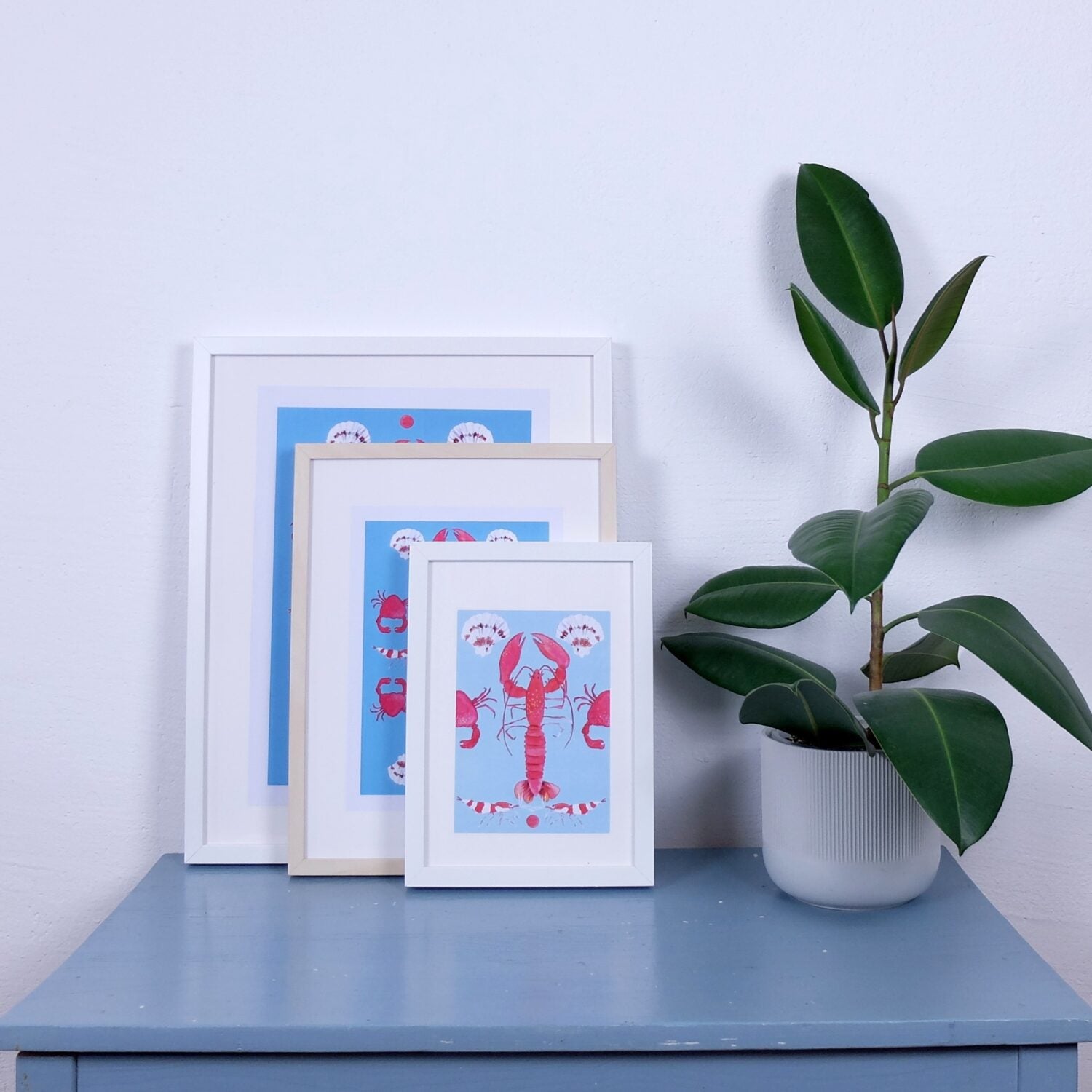 Drei gerahmte blaue Bilder mit Krabbenmotiven stehen auf einem Tisch neben einer grünen Pflanze in einem weißen Topf.