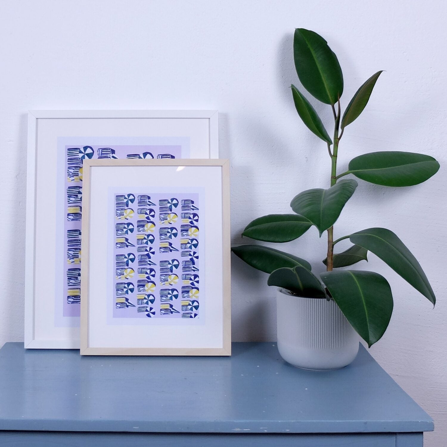 Zwei gerahmte Drucke mit blau-gelbem Muster stehen auf einem blauen Tisch neben einer Zimmerpflanze.