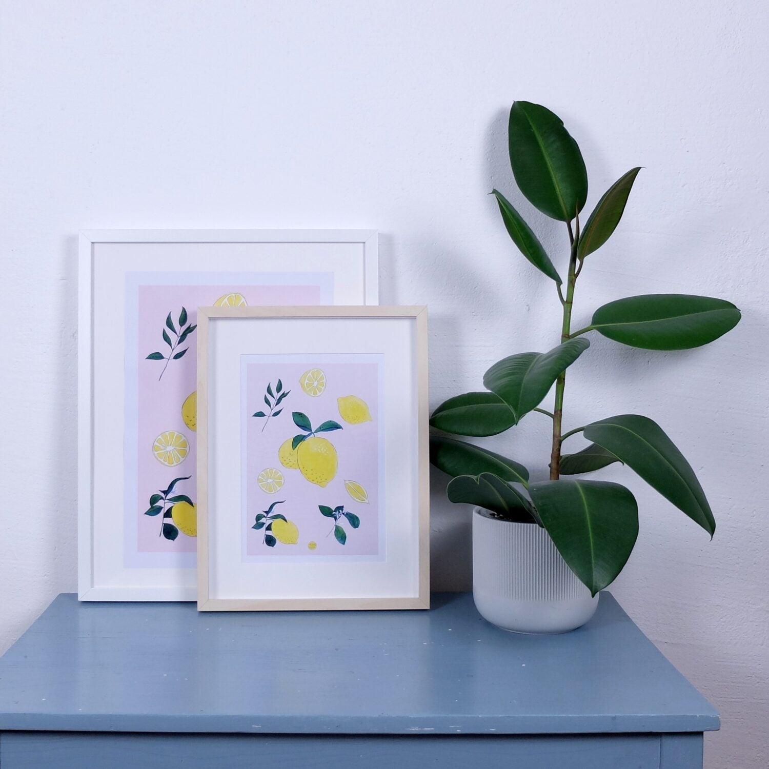 Zwei gerahmte Kunstwerke mit Zitronenmotiven auf einem blauen Tisch neben einer Topfpflanze.