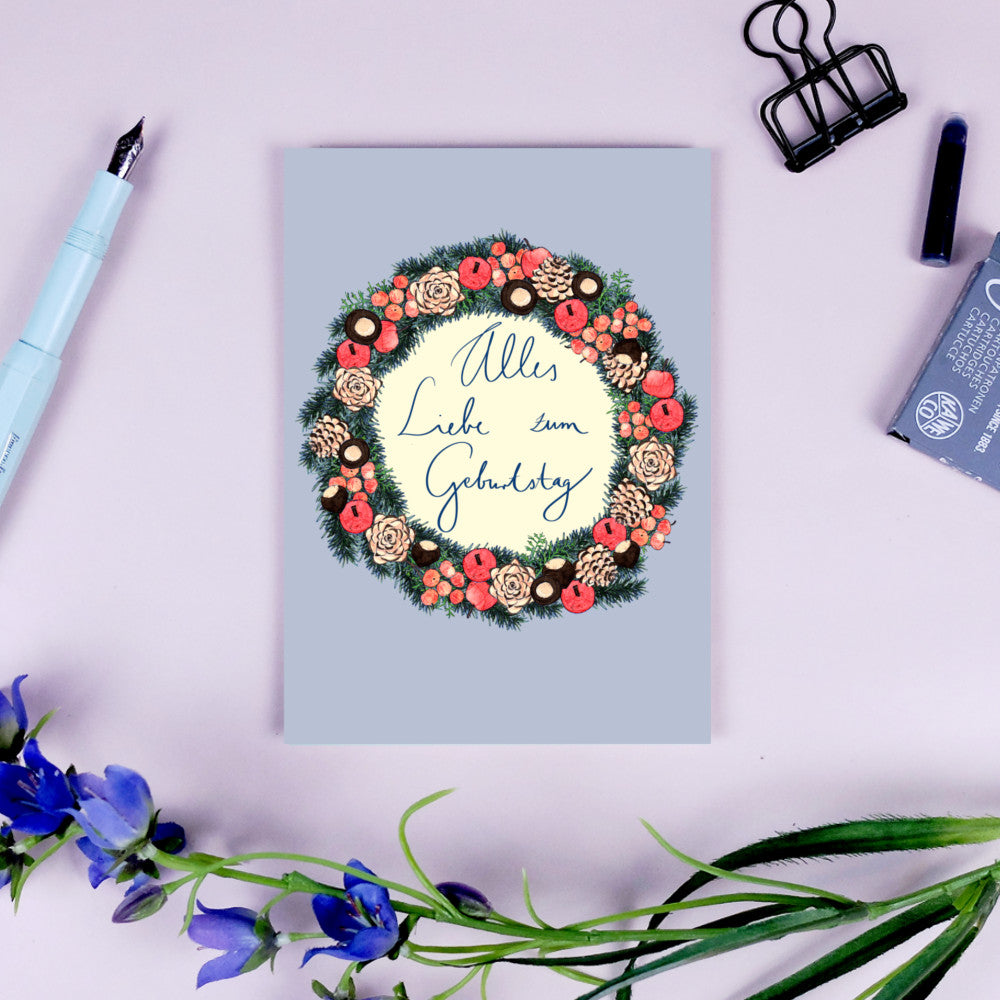 Geburtstagskarte mit Blumenkranz und der Aufschrift "Alles Liebe zum Geburtstag" auf einem Schreibtisch mit Dekoration.