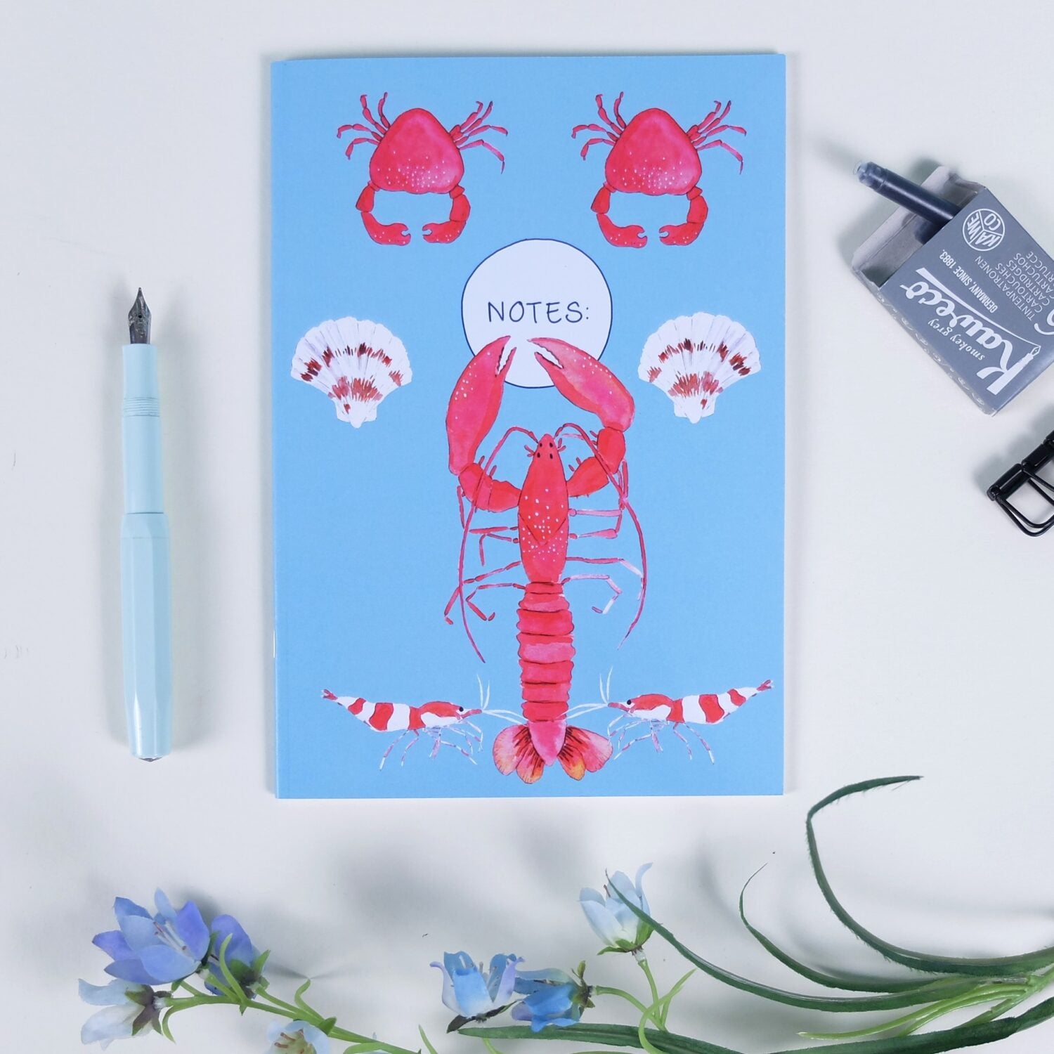 Blaues Notizbuch mit Hummer, Krabben und Muscheln, neben einem Füllfederhalter, Radiergummi und blauen Blumen.