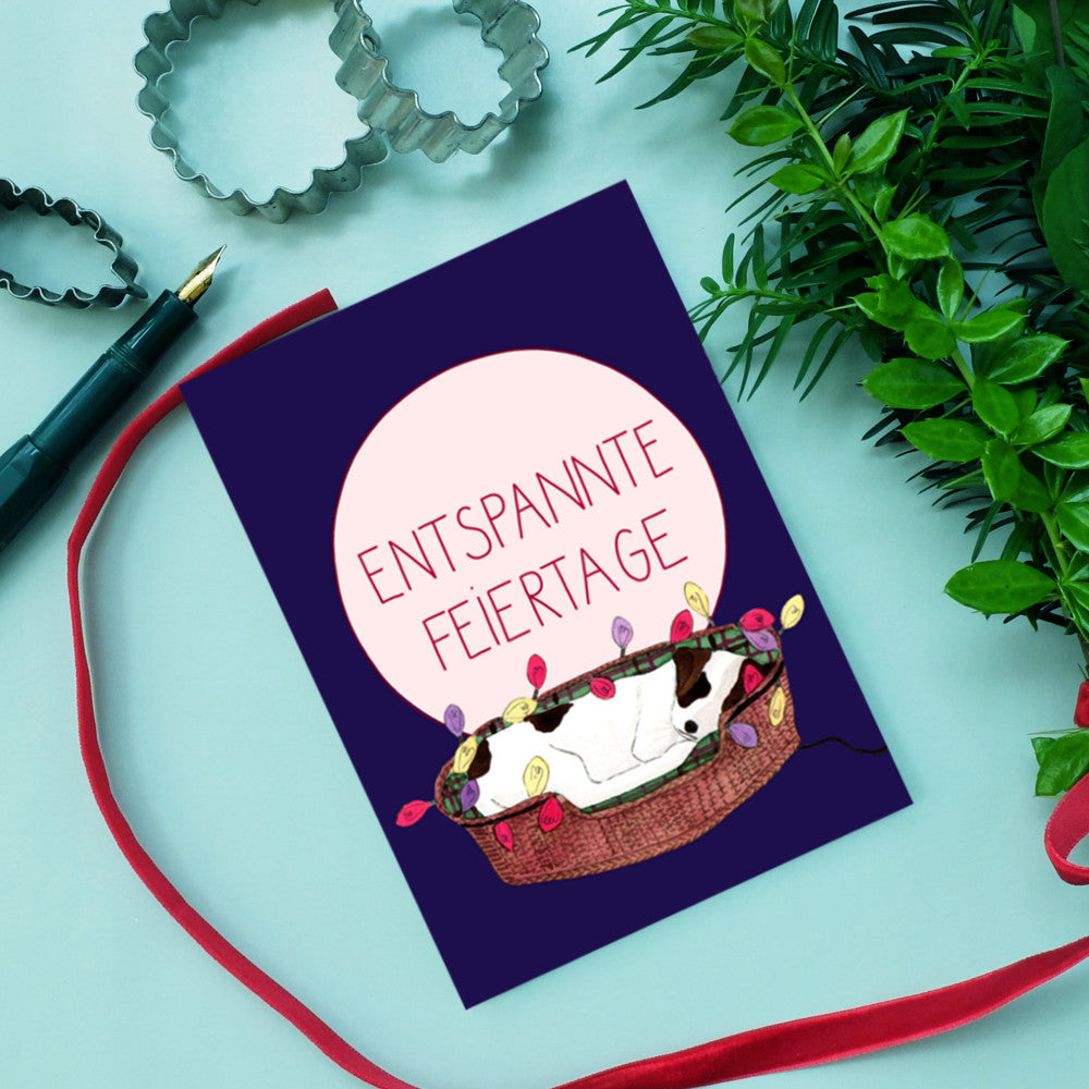 Eine Grußkarte auf blauem Hintergrund zeigt einen schlafenden Hund in einem Körbchen mit Lichterkette und der Aufschrift "Entspannte Feiertage".
