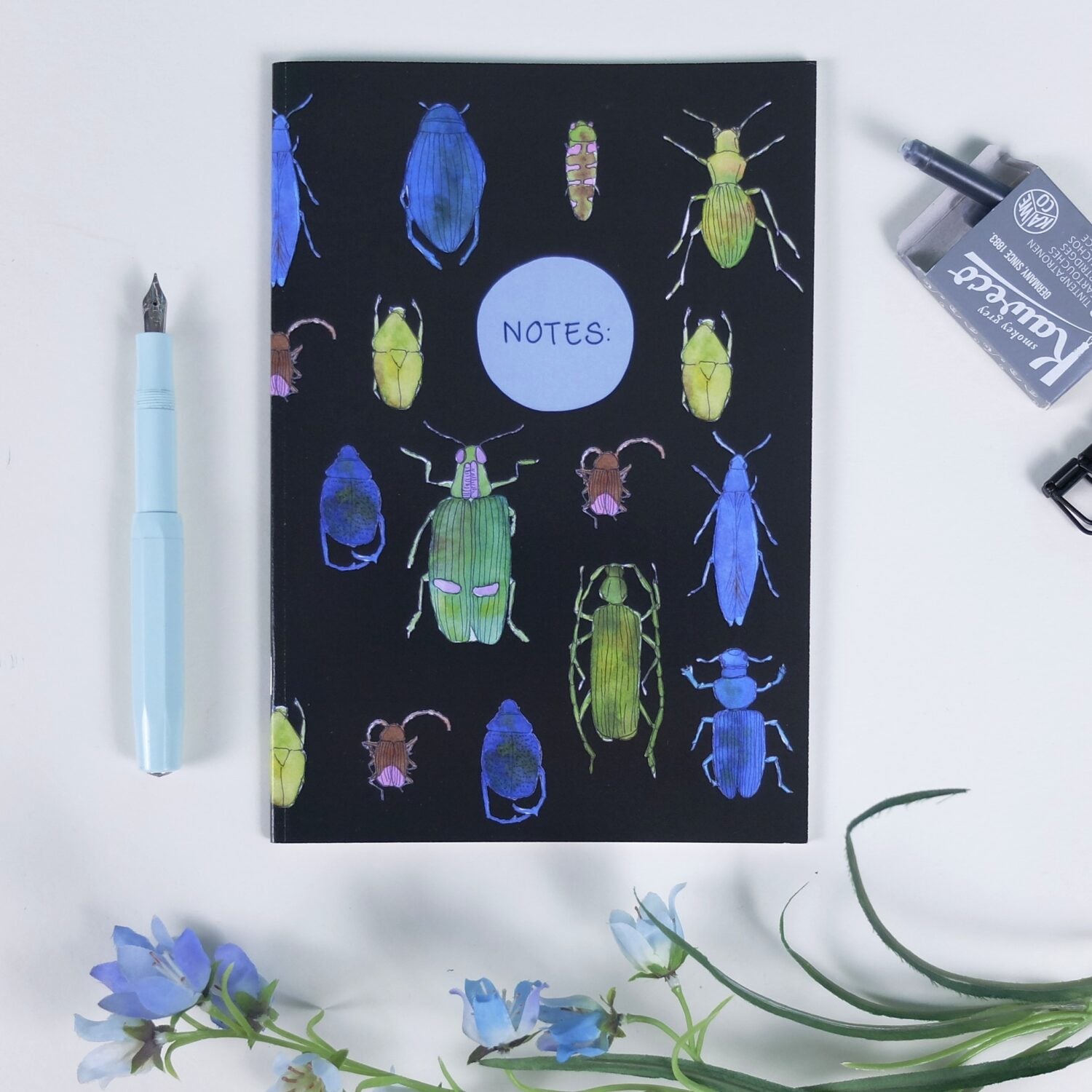 Notizbuch mit bunten Käfern und dem Wort "NOTES", daneben ein blauer Füller, eine blaue Blume und eine Tintenpatrone.