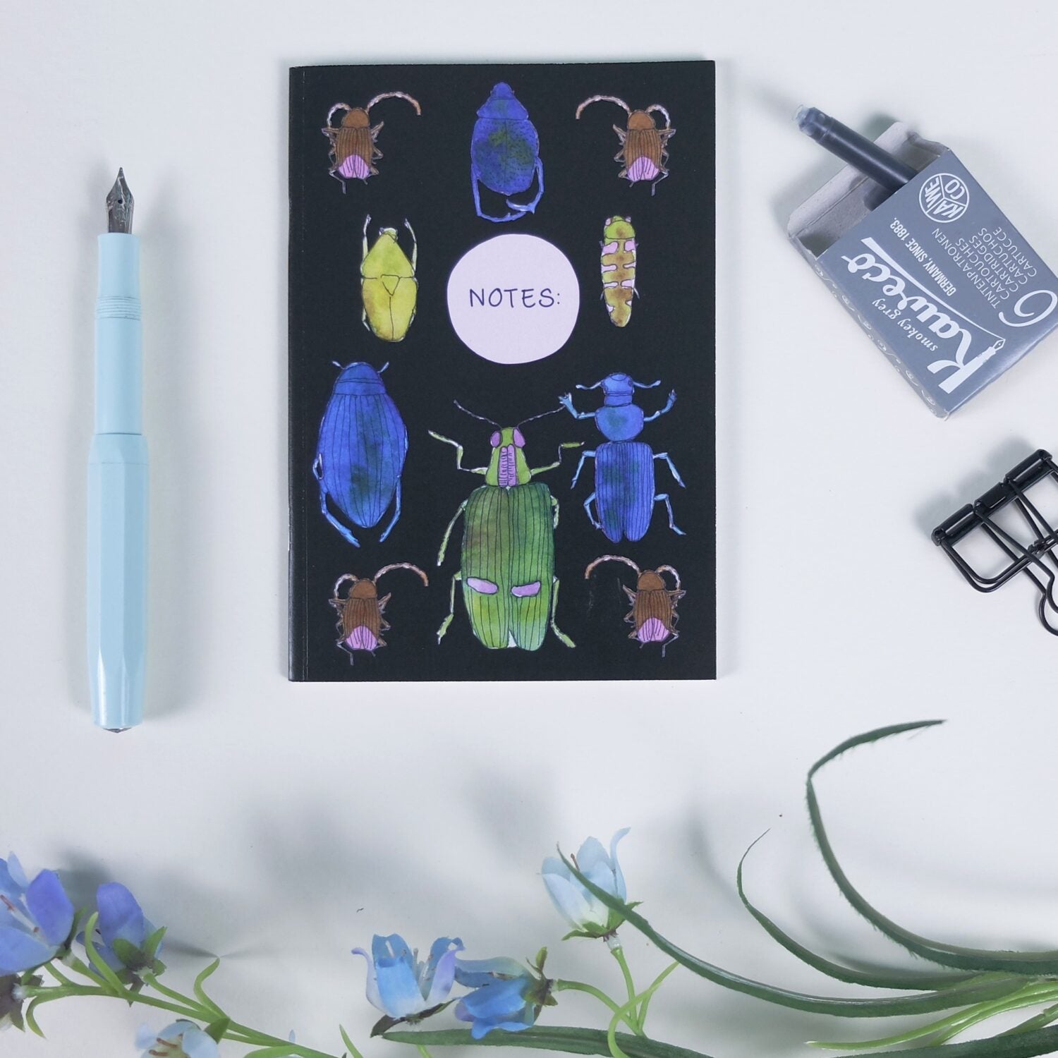 Notizbuch mit bunten Käfern, ein blauer Füller und Zubehör auf weißem Hintergrund mit blauen Blumen.