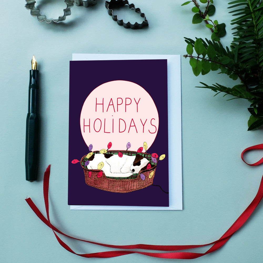 Karte mit "Happy Holidays"-Text, zeigt einen Hund im Korb mit Lichterketten. Kugelschreiber und Pflanzen im Hintergrund.