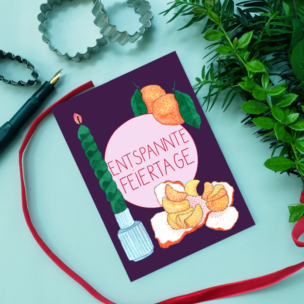 Eine Grußkarte mit der Aufschrift "Entspannte Feiertage" zeigt Mandarinen, eine Kerze und grüne Pflanzen.