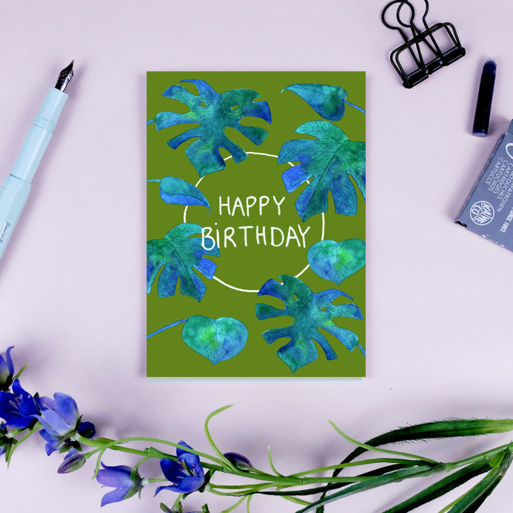 Geburtstagskarte mit blauen Blättern auf grünem Hintergrund, "Happy Birthday" in der Mitte; daneben Blumen und Stift.