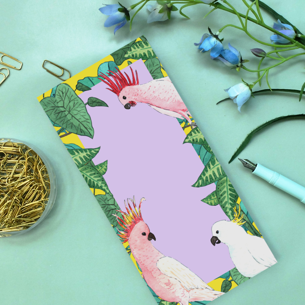 Notizblock mit Papageien-Illustrationen, umgeben von Papierclips, einem hellblauen Stift und blauen Blumen.