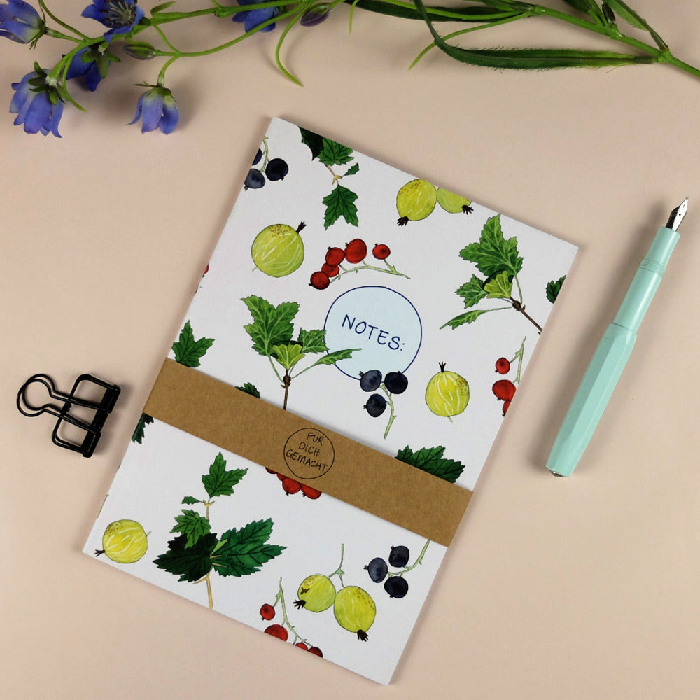 Notizbuch mit Frucht- und Blattdesign, ein hellblauer Füller und eine schwarze Klammer auf beigem Hintergrund.