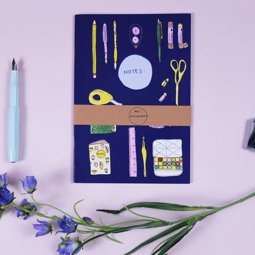 Notizheft mit bunten Zeichnungen von Schreibwaren auf dem Cover, daneben ein Füller und Blumen.