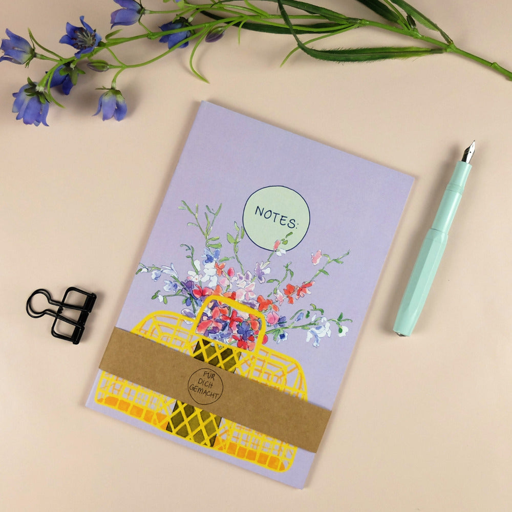 Notizbuch mit Blumendruck, dazu ein mintgrüner Füllfederhalter, eine schwarze Büroklammer und lila Blüten.