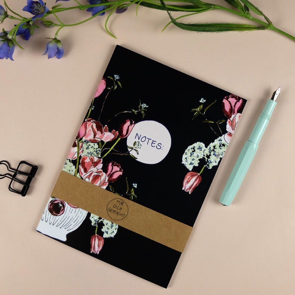 Schwarzes Notizbuch mit Blumendesign, daneben ein türkiser Füllfederhalter und eine Büroklammer auf beige Hintergrund.