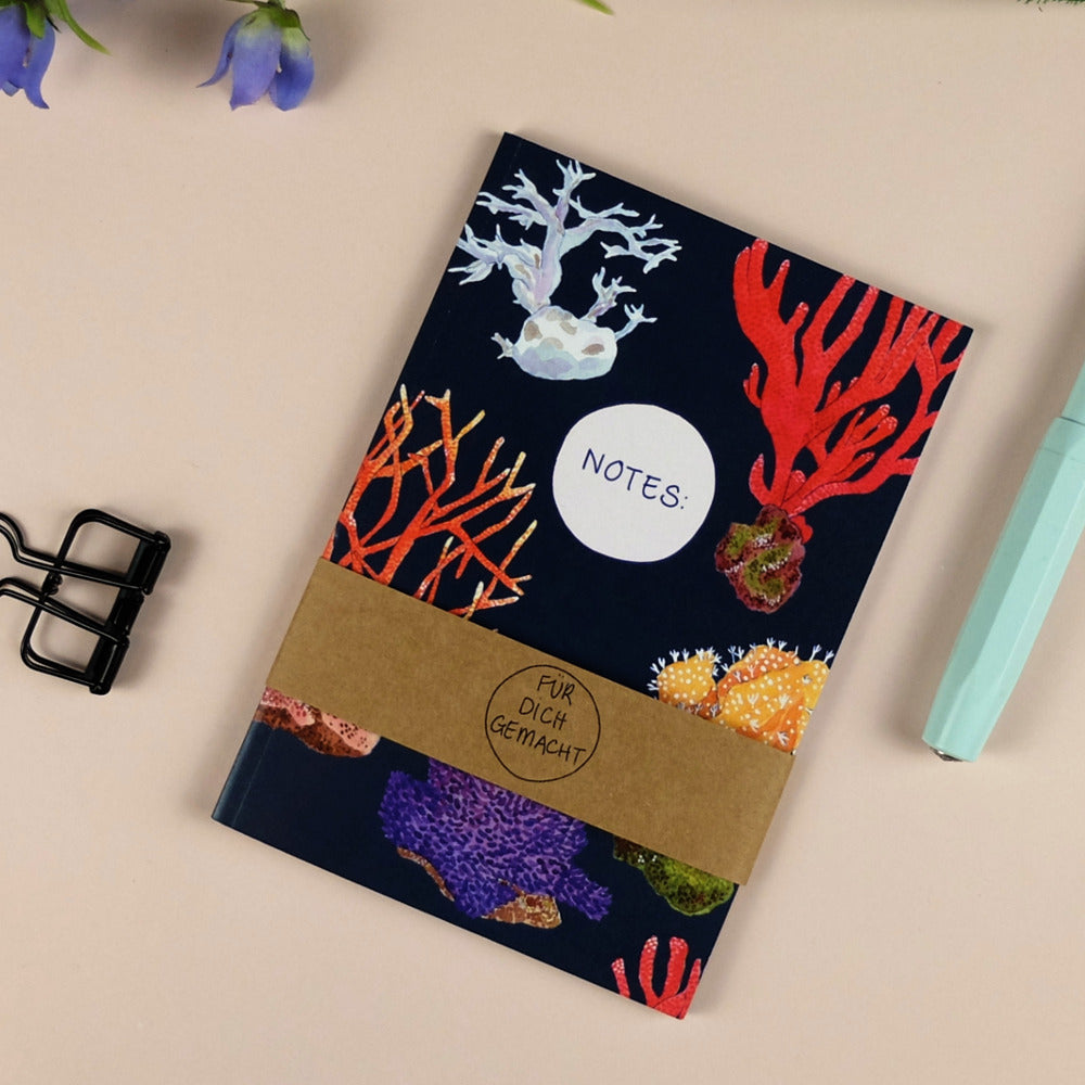Notizbuch mit Korallenmuster auf dunkelblauem Hintergrund, umgeben von einer Büroklammer und einem türkisen Stift.