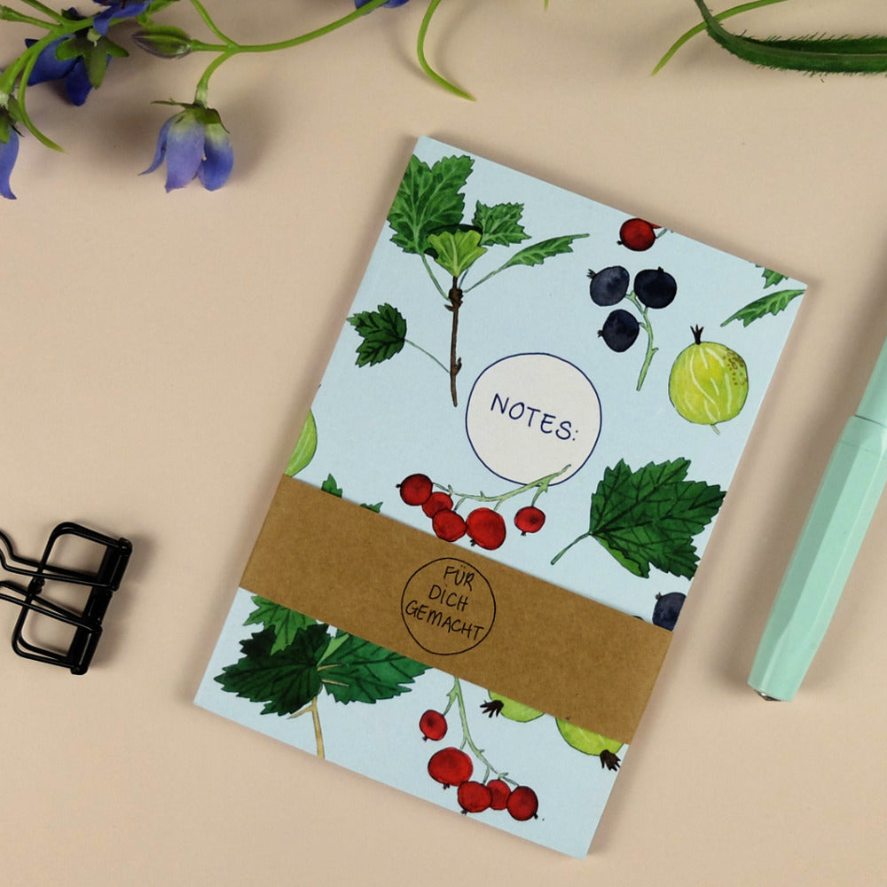 Notizheft mit Beeren- und Blattmuster auf einem Tisch, daneben ein Stift und eine Büroklammer.