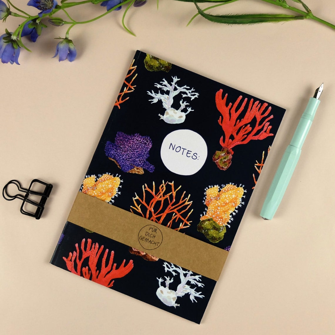 Notizbuch mit Korallenmotiv, Stift und Büroklammer auf beigem Hintergrund; Aufschrift "Notes" und "Für dich gemacht".