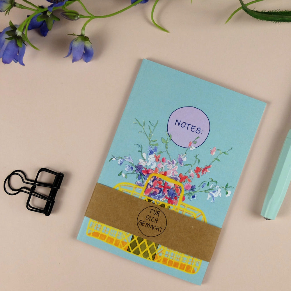 Notizbuch mit Blumenmotiv, Aufschrift "NOTES:", Stiftehalter, und ein Stift auf beigem Hintergrund.