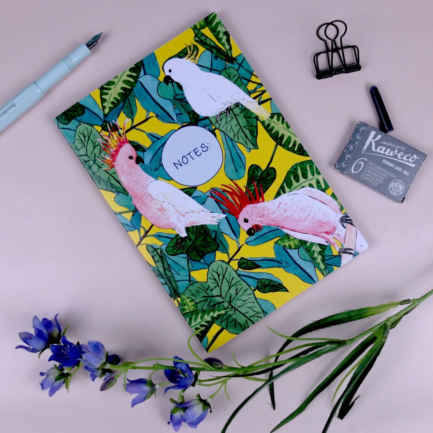 Notizbuch mit buntem Papageien-Muster, Feder, Blumen, Tintenpatronen und einer Klammer auf rosa Hintergrund.
