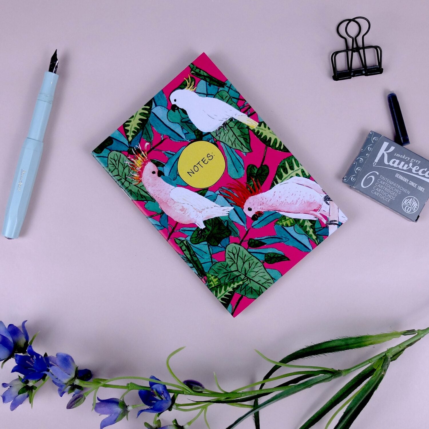 Buntes Notizbuch mit Papageienmotiv, blauer Füller, Klammern, Tintenpatronen und lilafarbene Blumen auf rosa Hintergrund.
