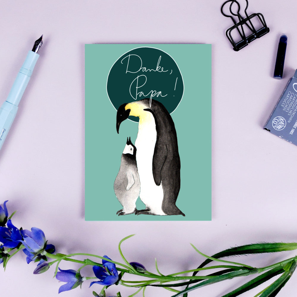 Karte mit Pinguin-Eltern und Küken, sowie dem Text „Danke, Papa!“ auf hellgrünem Hintergrund, umgeben von Schreibwaren.