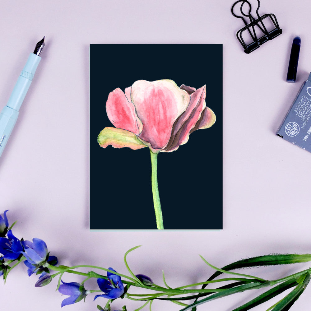 Karte mit rosa Blume auf dunklem Hintergrund, umgeben von einem Stift, Klammer und blauen Blumen.