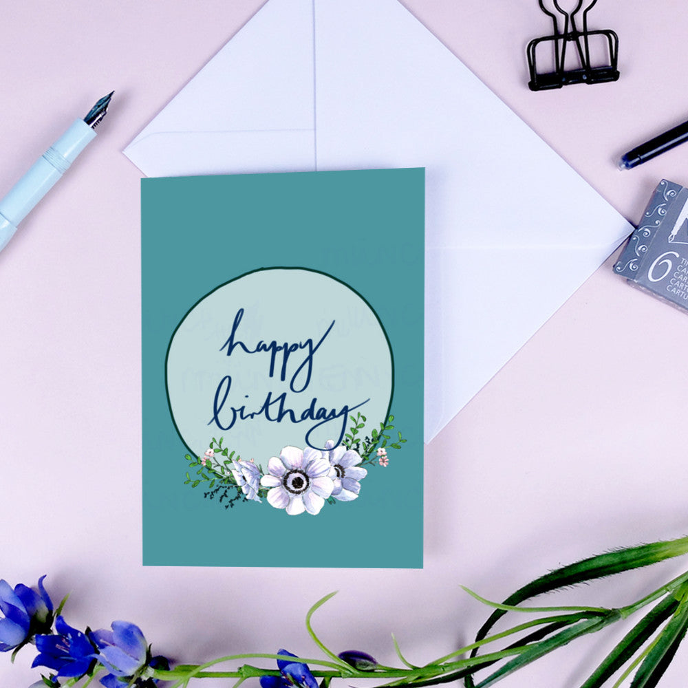 Geburtstagskarte mit Schriftzug "Happy Birthday" und Blumenmotiv, umgeben von einem Stift und Blumen auf einem Tisch.