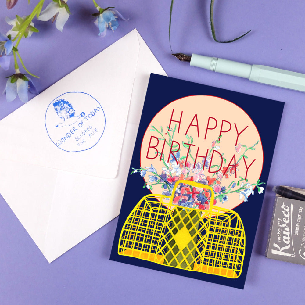 Geburtstagskarte mit "Happy Birthday" und Blumenmotiv neben Briefumschlag und Stift auf lila Hintergrund.