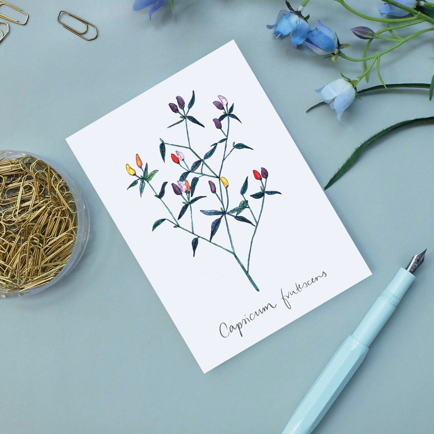 Karte mit einer handgezeichneten Pflanze auf einem Schreibtisch mit Büroklammern, Blumen und einem Stift.