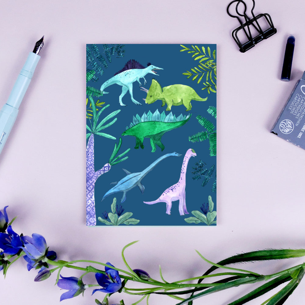 Bunte Dinosaurier-Illustration auf blauem Hintergrund, umgeben von Pflanzen, neben Stift und Blumen auf einem Tisch.