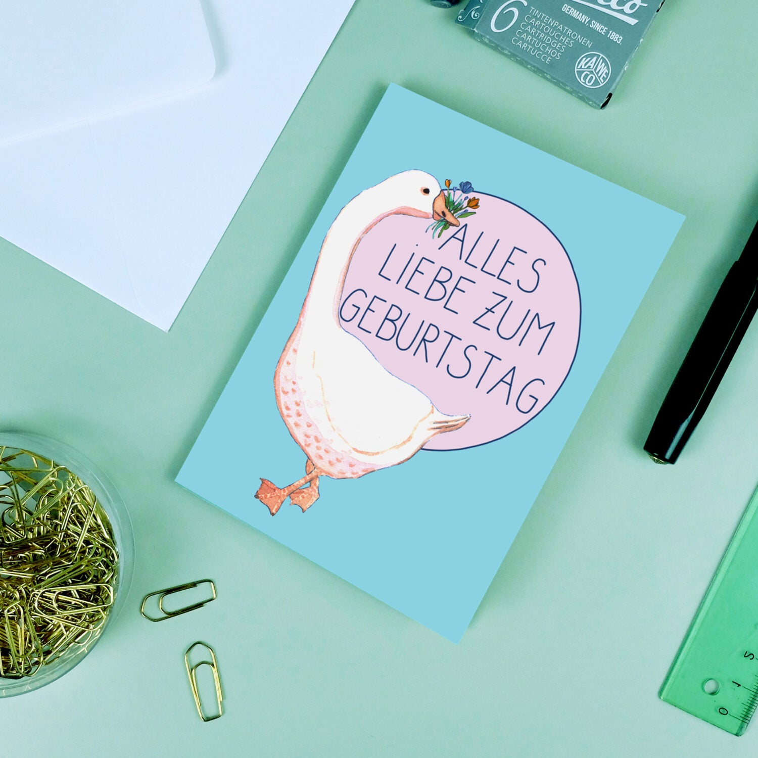 Geburtstagskarte mit einer Ente, die Blumen hält und dem Text "Alles Liebe zum Geburtstag" auf einem grünen Tisch.