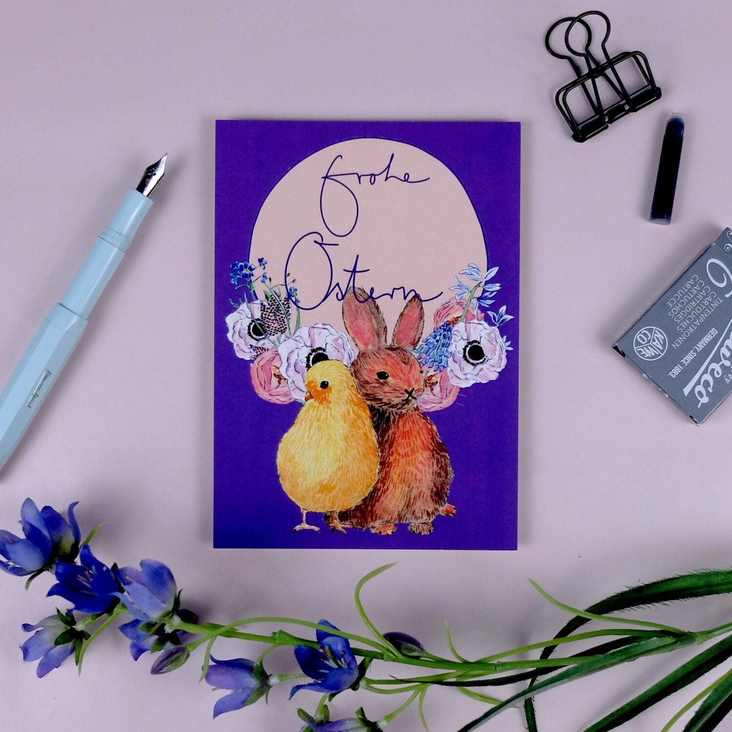 Auf einer Karte steht "Frohe Ostern" mit einem Küken und Hasen vor Blumen. Daneben liegen Stift, Klammer und Blumen.