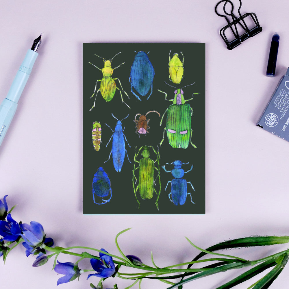 Postkarte mit bunten Käferillustrationen, umgeben von blauen Blumen, Stift, Büroklammer und Tintenkapsel.