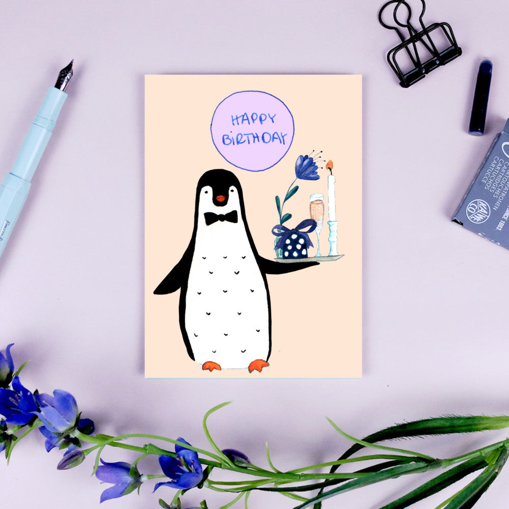 Eine Geburtstagskarte mit einem gezeichneten Pinguin, der ein Tablett mit Kerzen und einem Geschenk hält, umgeben von Blumen.