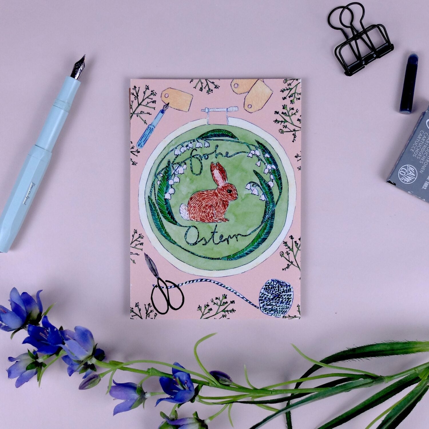 Grußkarte mit Hase und "Frohe Ostern"-Text, umgeben von Blumen, neben einem Füllfederhalter und blauen Blumen.