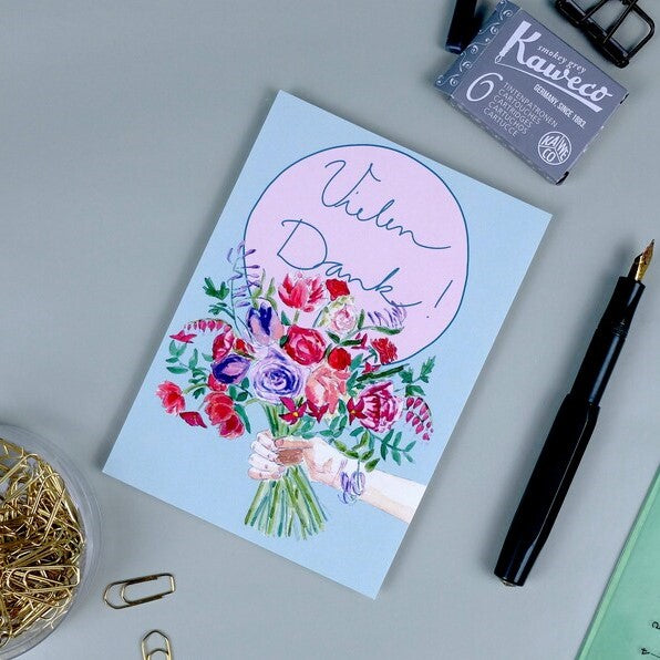 Grußkarte mit handgemaltem Blumenstrauß und der Aufschrift "Vielen Dank!", daneben Füller und Büroklammern.