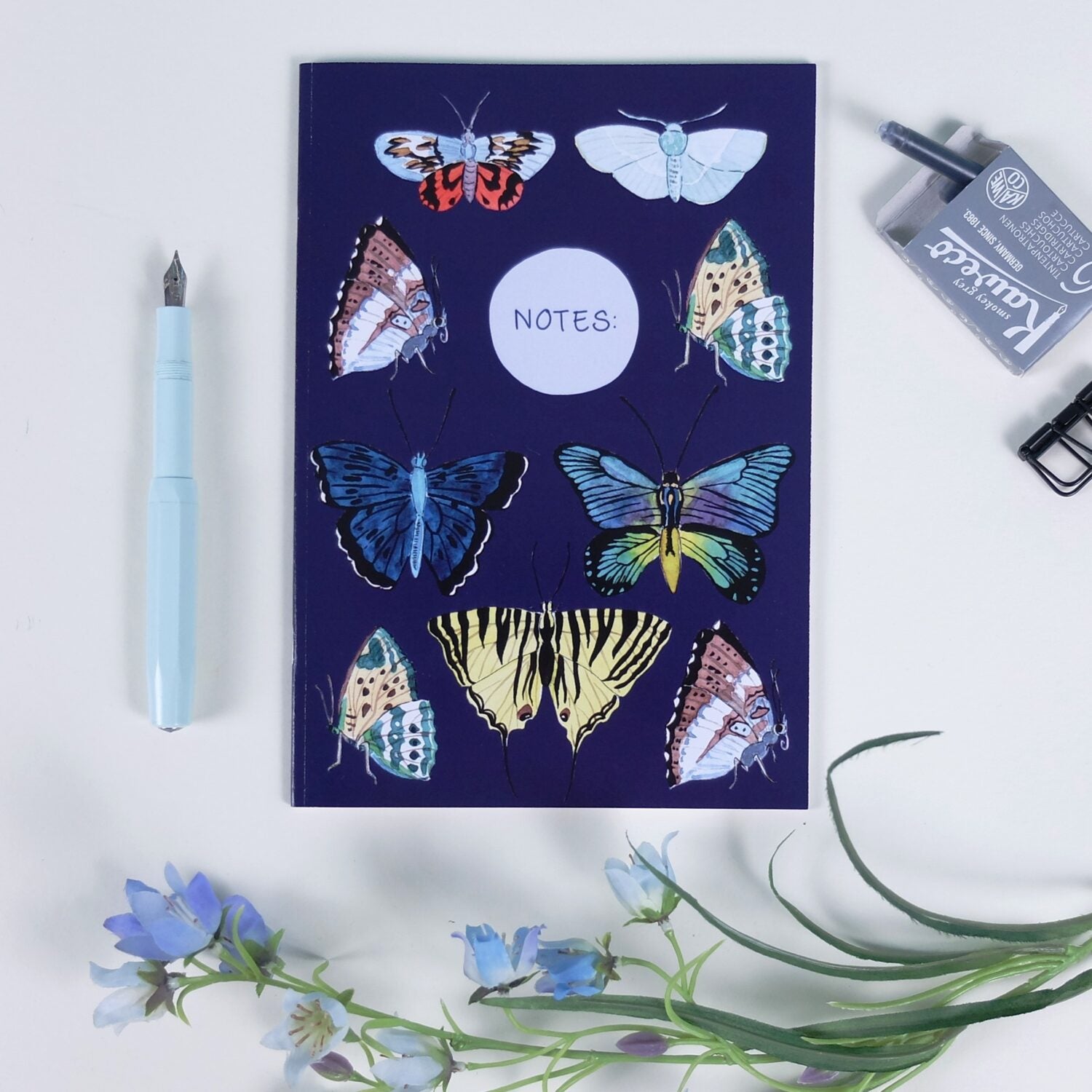 Notizbuch mit bunten Schmetterlingsmotiven, daneben ein blauer Füller und Blumen auf weißem Hintergrund.