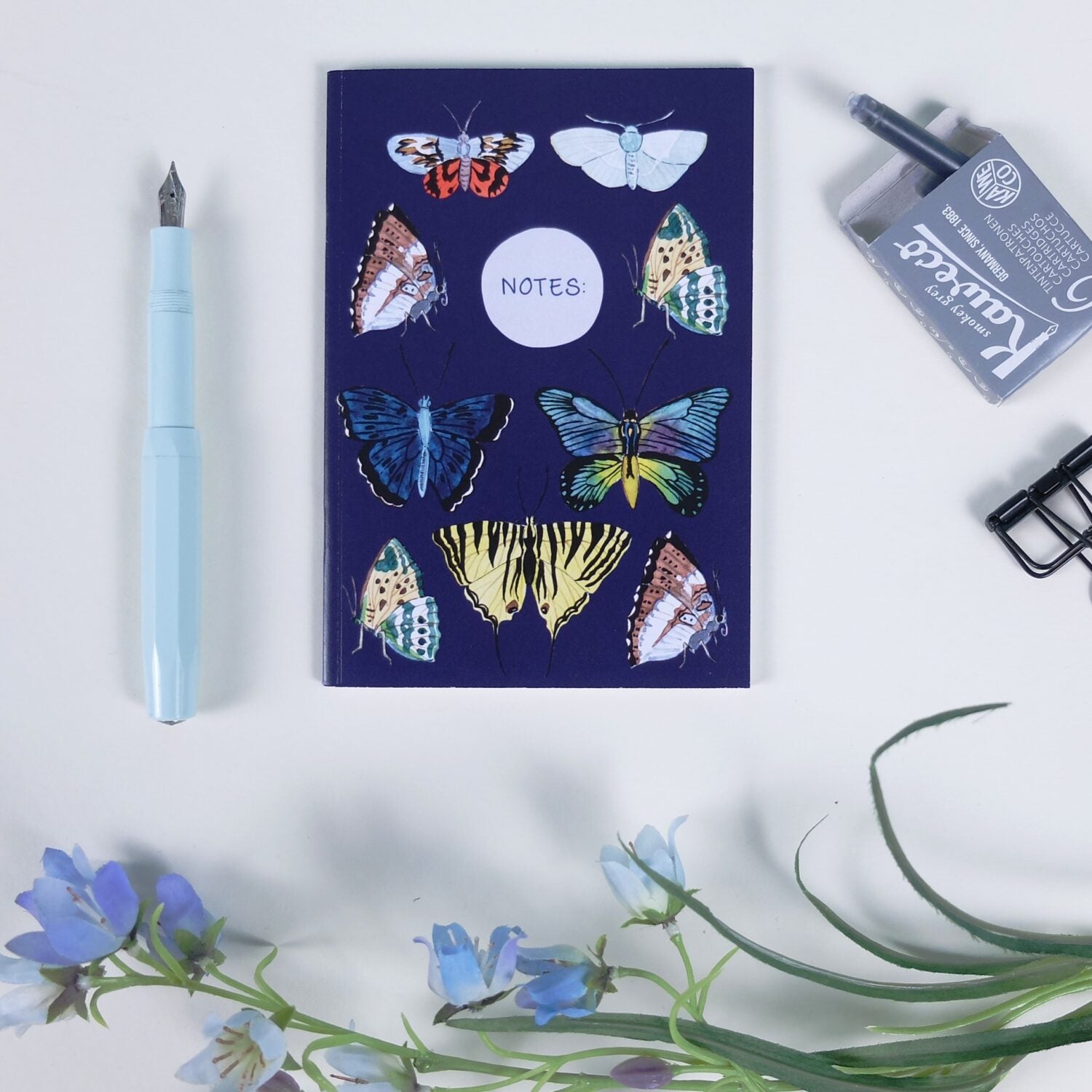 Notizbuch mit Schmetterlingen auf dem Cover, umgeben von einem blauen Stift, Tinte und blauen Blüten auf einem weißen Hintergrund.