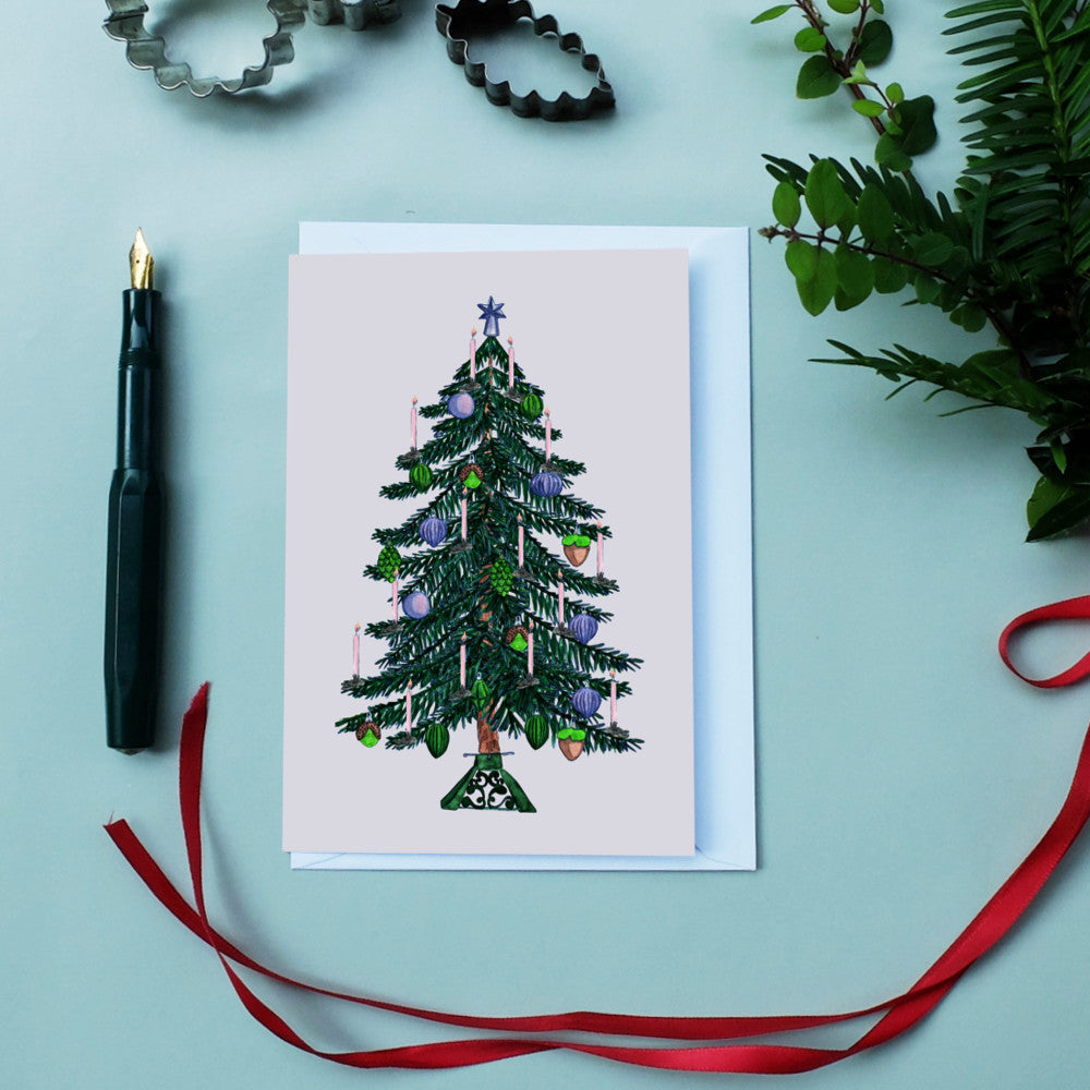 Weihnachtskarte mit geschmücktem Tannenbaum, umgeben von einem Stift, Backformen und einem roten Band.