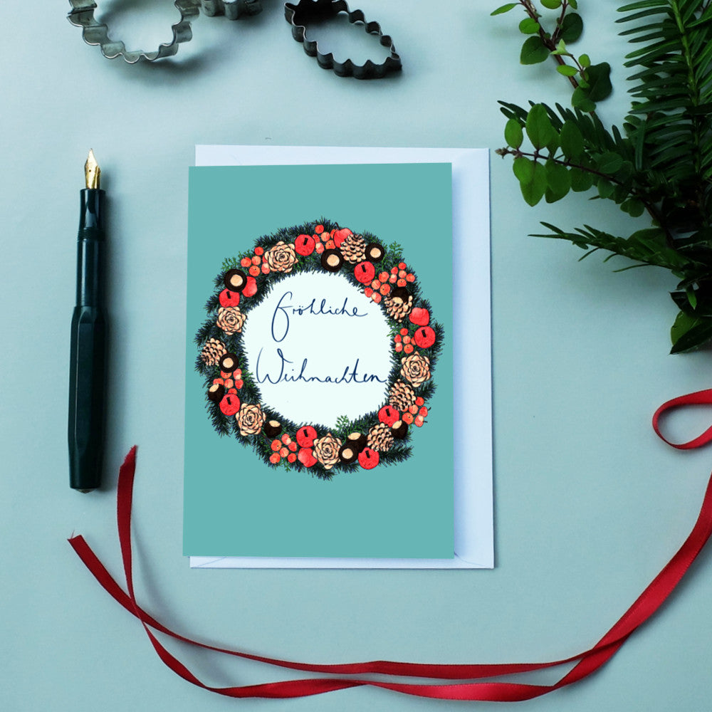 Weihnachtskarte mit Kranz und "Fröhliche Weihnachten"-Text, daneben ein Füllfederhalter und rote Schleife.