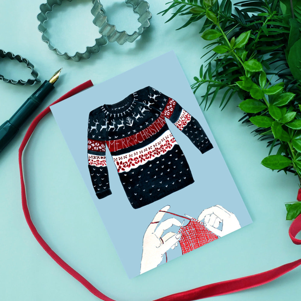 Weihnachtliche Grußkarte mit Pullovermotiv, Stricknadeln und Pflanzen auf einem mintgrünen Hintergrund.