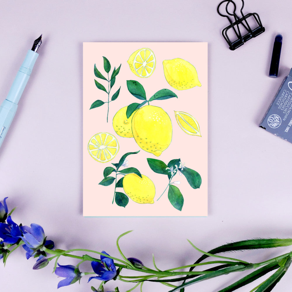 Illustration von Zitronen und Blättern auf einer Postkarte, umgeben von Blumen, einer Klammer und einem Stift.