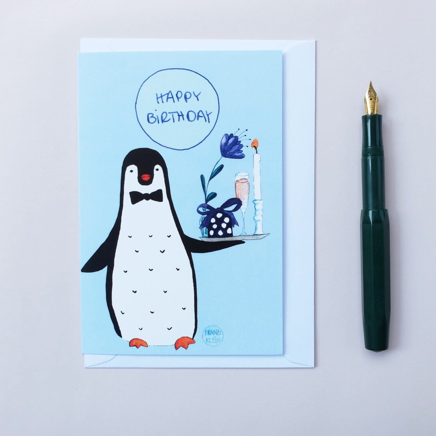 Grußkarte mit einem gezeichneten Pinguin, der eine Torte und eine Kerze hält, neben einem Füllfederhalter.