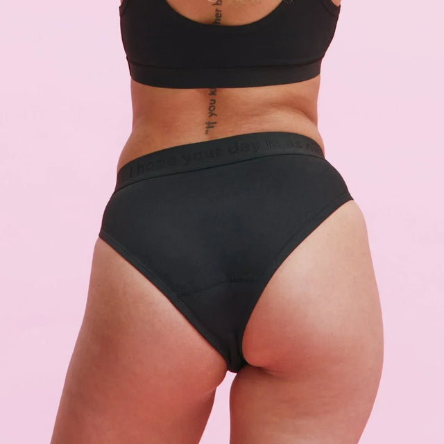 Eine Person mit langen blonden Haaren wird von hinten gezeigt, die einen schwarzen BH und schwarze Unterwäsche vor einem hellrosa Hintergrund trägt. Entlang ihrer Wirbelsäule ist eine Tätowierung zu sehen.