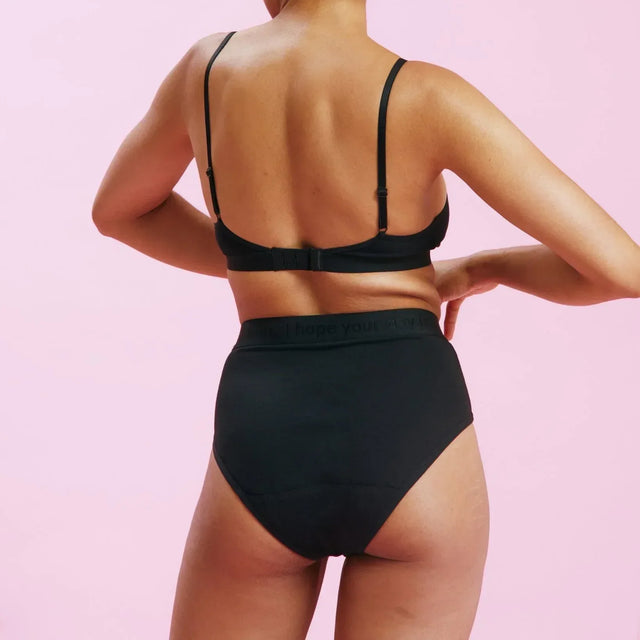 Eine Frau mit hochgestecktem Haar, von hinten gesehen, steht vor einem rosa Hintergrund und trägt einen schwarzen BH und schwarze Unterwäsche mit hoher Taille.