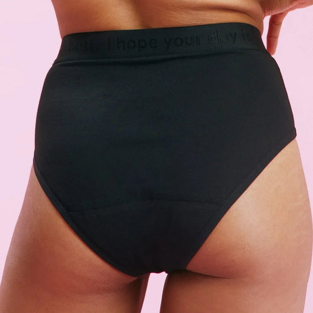 Eine Person, die schwarze Unterwäsche mit hoher Taille und einen passenden schwarzen BH trägt, steht vor einem hellrosa Hintergrund, der von hinten und von der Taille abwärts gezeigt wird. Der Bund der Unterwäsche ist mit einem dezenten Text versehen.