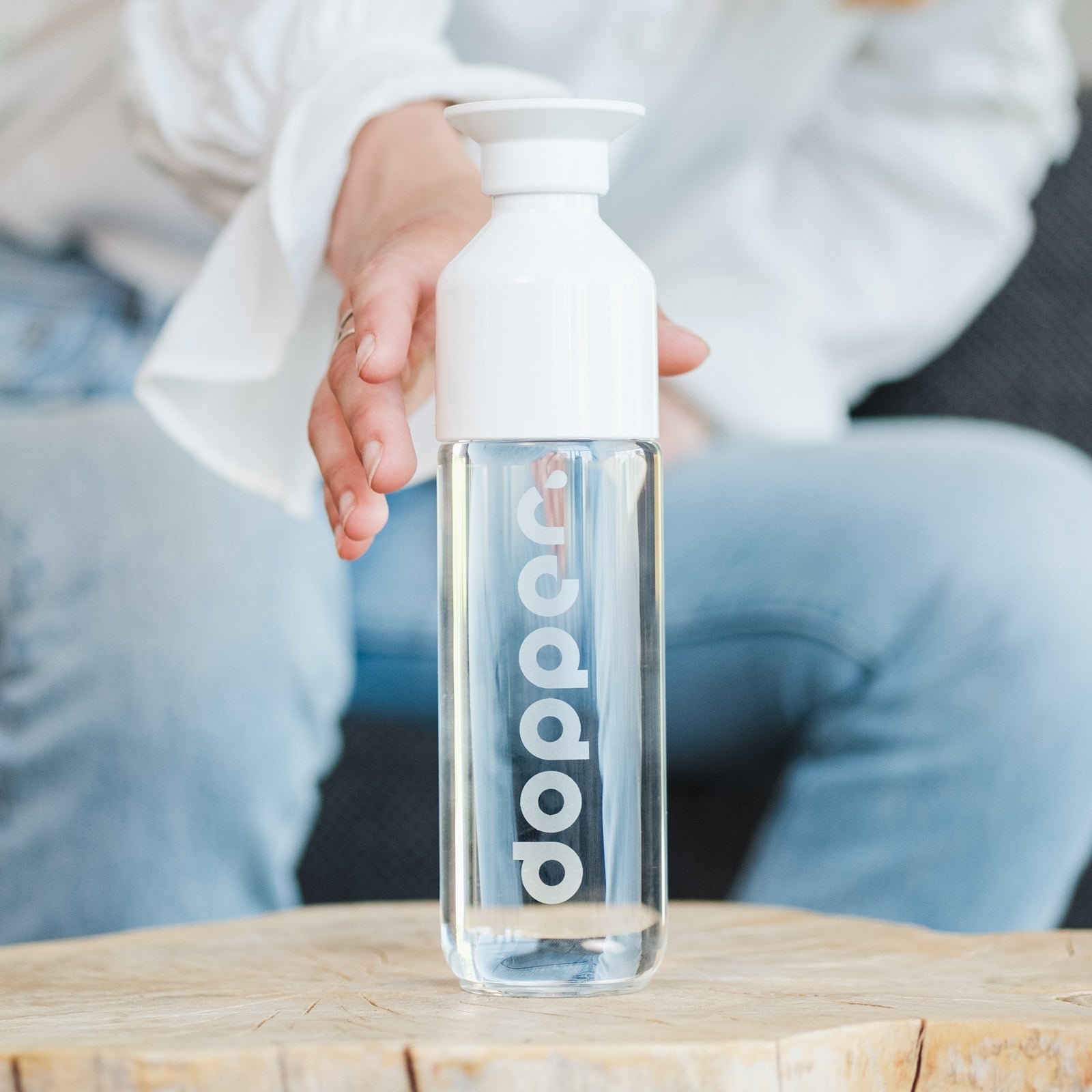 Hand greift nach einer transparenten Dopper-Wasserflasche mit weißem Deckel auf einem Holztisch.