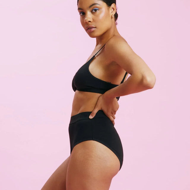 Eine Frau mit hochgestecktem Haar posiert in schwarzer Unterwäsche vor einem einfarbigen rosa Hintergrund und blickt selbstbewusst in die Kamera.