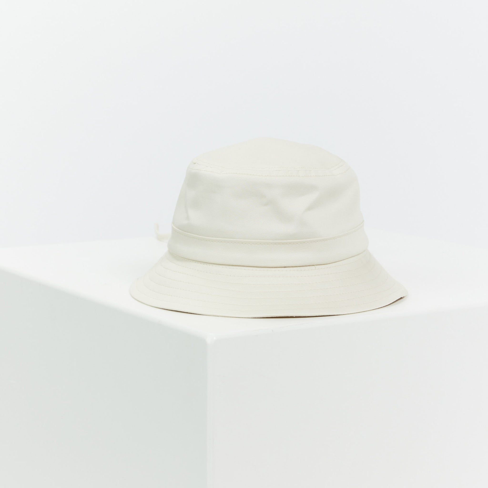 Weißer Fischerhut auf einem weißen, quaderförmigen Podest in minimalistischem Stil.